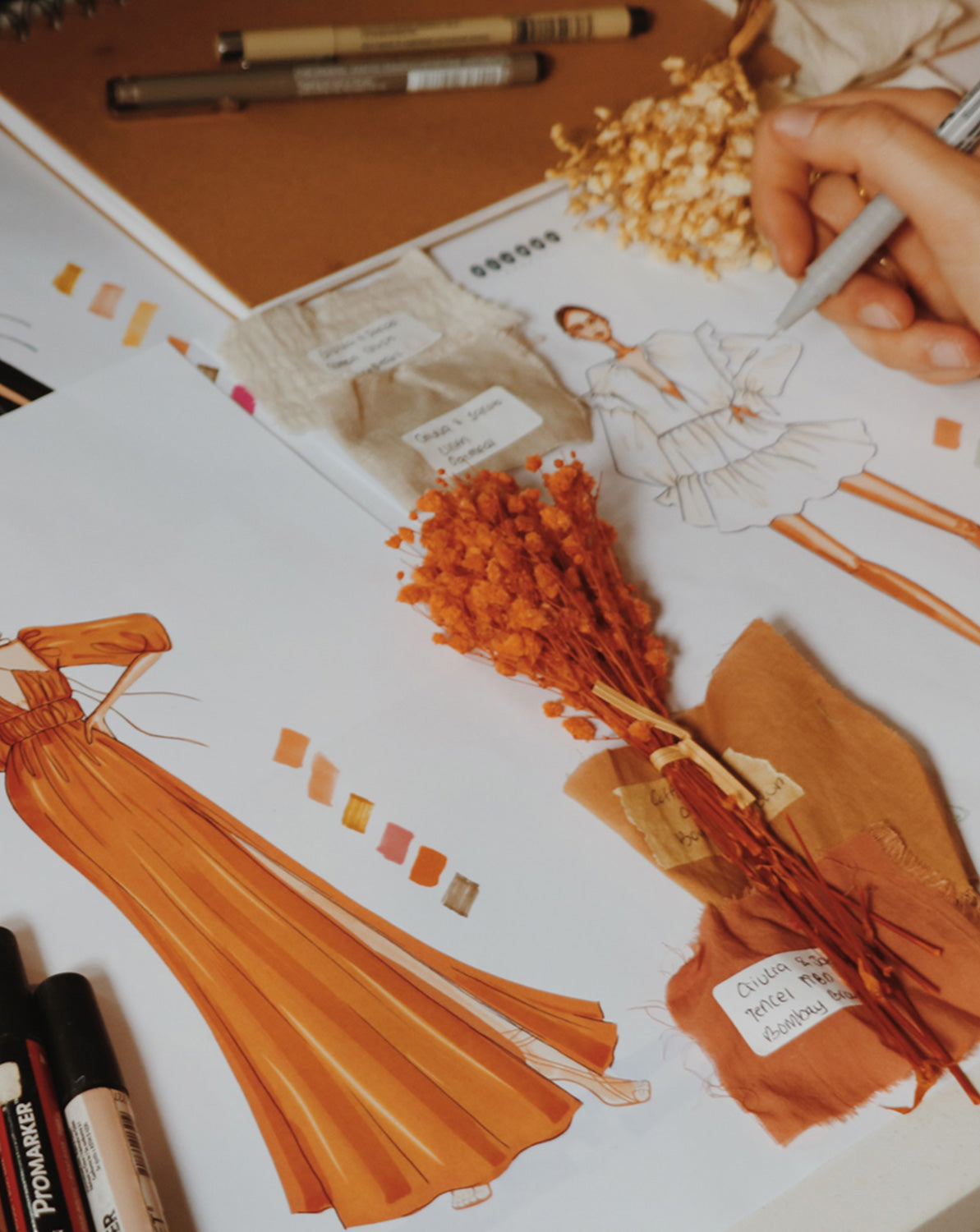 Close-up of fashion sketches of sustainable and ethical fashion, one maxi rust dress and one mini white dress, with dried flowers and markers around the drawings.