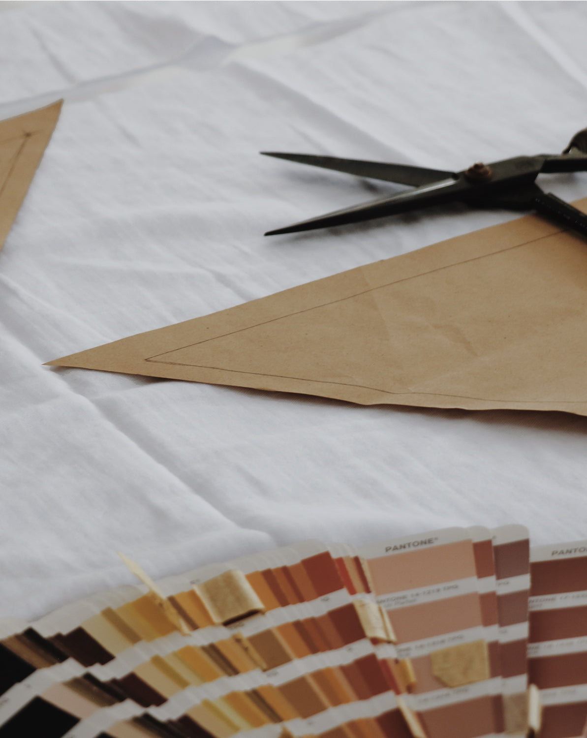 Close-up of a pair of tailor scissors on top of a white piece of cotton fabric, with kraft paper sewing pattern and pantone colour swatches.