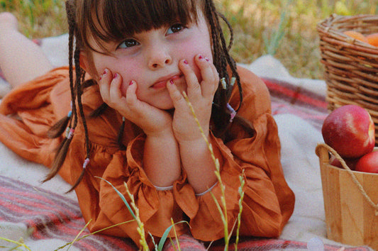 Toddler wearing brown shiny dress with long puff sleeves, laying on a picnic rug, in a fruit orchard, with baskets full of fruits.