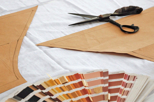 Close-up of sewing scissors, white fabric, brown pattern paper and colour swatches on a sewing table.