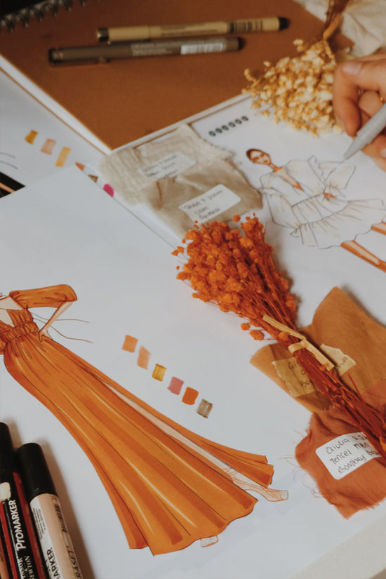 Close-up of photograph of fashion sketches of a terracotta maxi dress and a white mini dress, with fabric swatches and orange dried flowers on the dress and 2 markers around the drawings.