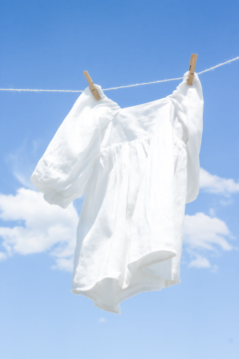 White breathable natural linen dress for girls by Fiamma Studio, hanging on the string with blue sky in the background.