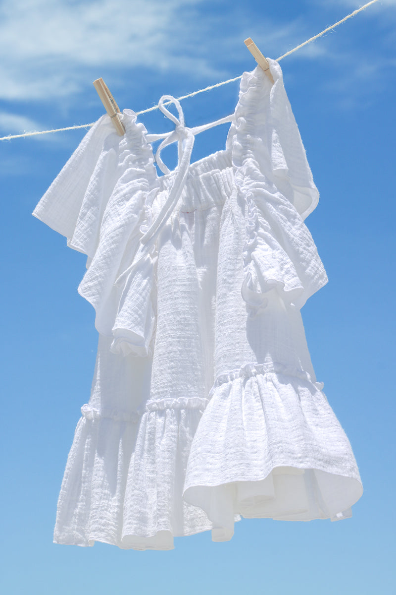 White cotton gauze tiered dress for girls, with flowy frills sleeves and bottom hem flounce. The dress is hanging on the clothesline, with blue cloudy sky in the background.