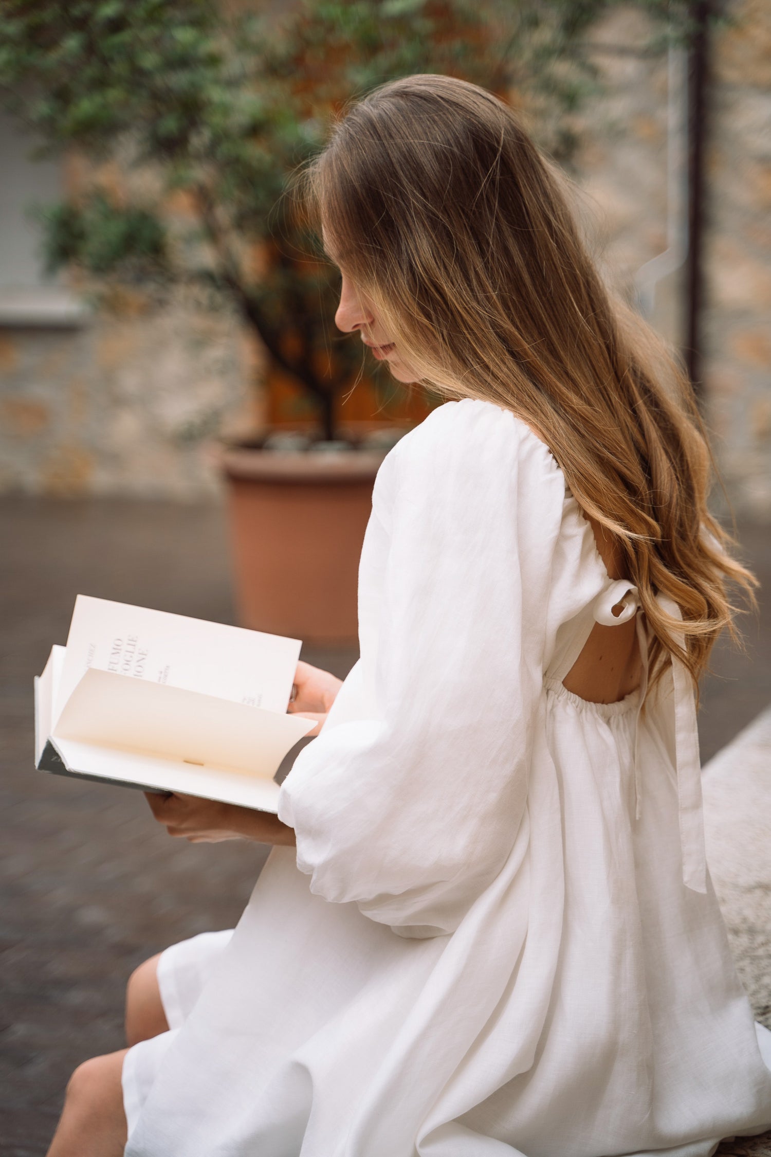 Woman sitting while reading a book, wearing white sustainable linen Fiamma Studio mini dress, with open back and bow ties closure.