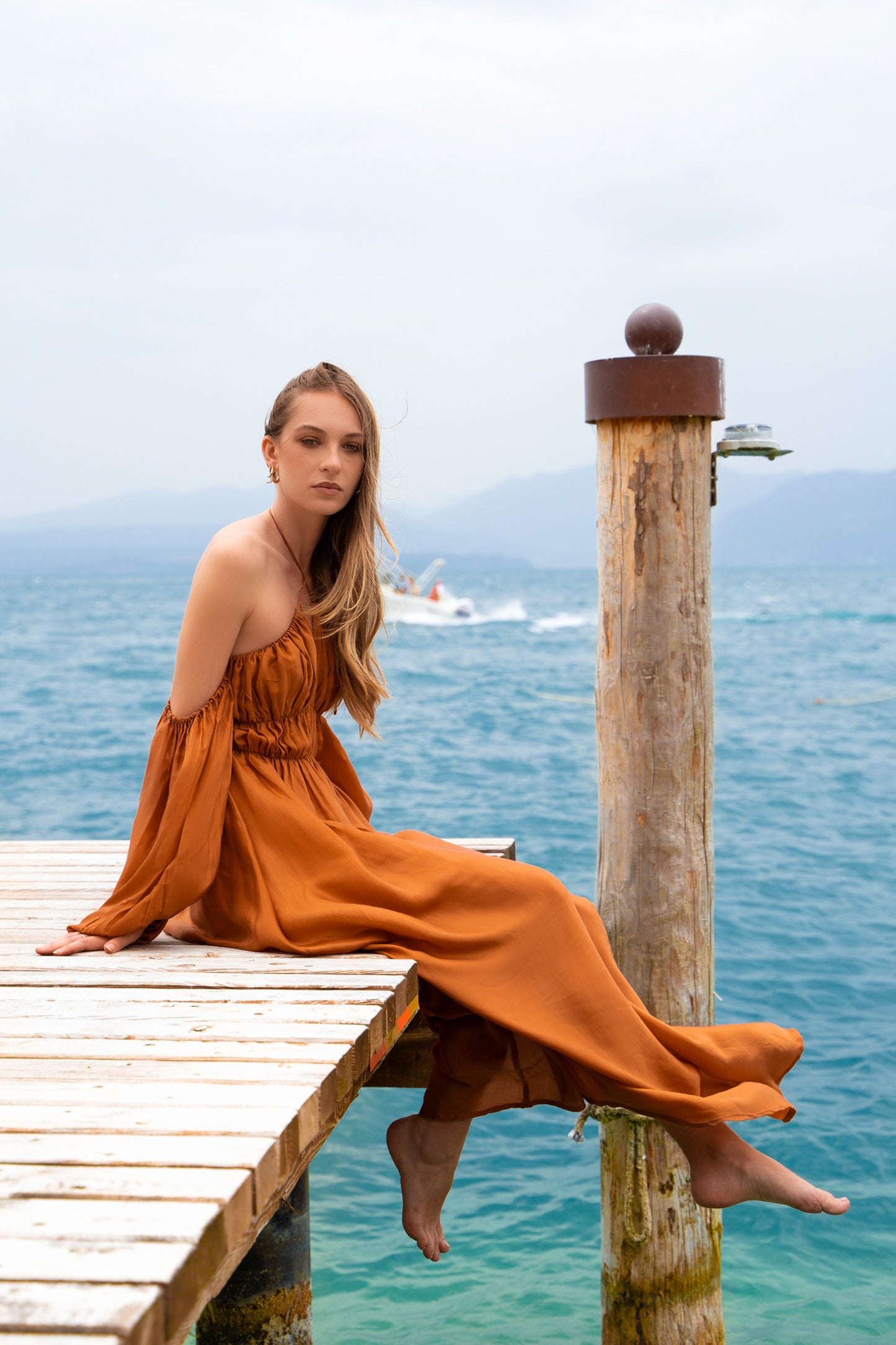 Woman sitting on a lake pier, wearing a terracotta maxi dress, with puff sleeves, shirred bust, a-line maxi skirt skirt.