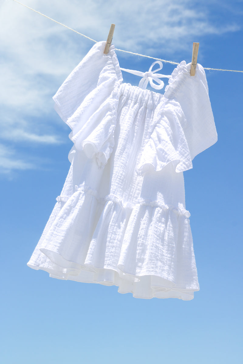 Entire figure of white cotton gauze tiered dress for girls, with flowy frills sleeves and bottom hem flounce. Textured fabric, natural and sustainable clothing. The dress is hanging on the clothesline and the background is cloudy blue sky.