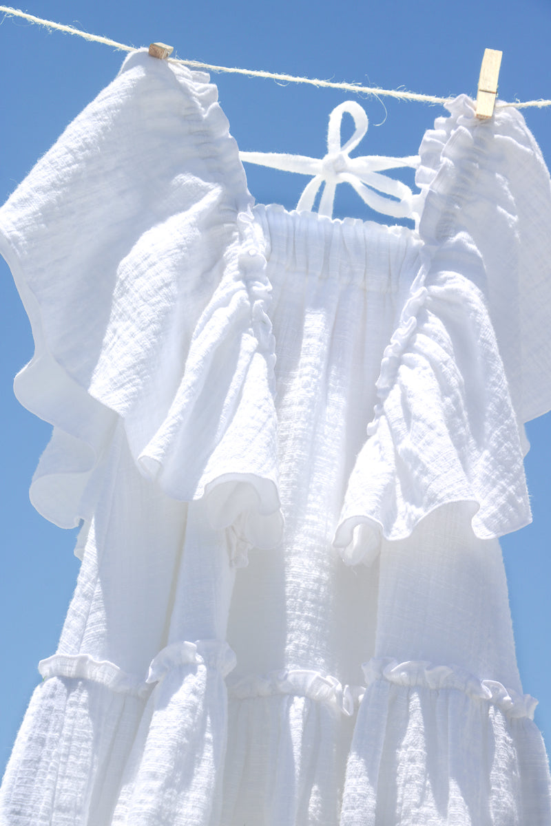 Close-up of white cotton gauze tiered dress for girls, with flowy frills sleeves and bottom hem flounce. Textured fabric, natural and sustainable clothing. The dress is hanging on the clothesline and the background is cloudy blue sky.