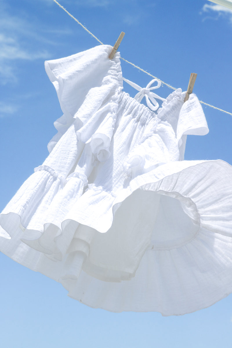 White cotton gauze tiered dress for girls, with flowy frills sleeves and bottom hem flounce. Textured fabric, natural and sustainable clothing. The dress is hanging on the clothesline and the background is cloudy blue sky.